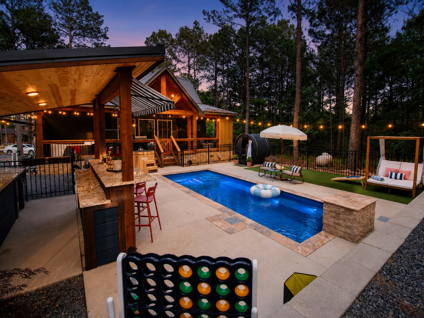 Gilded Spur cabin backyard hero at sunset, Hochatown OK luxury vacation rental: in-ground pool, covered pool bar with green-and-white striped awning, cabin lit up, cornhole with branded throw pillows, fire pit, hammocks, string lights