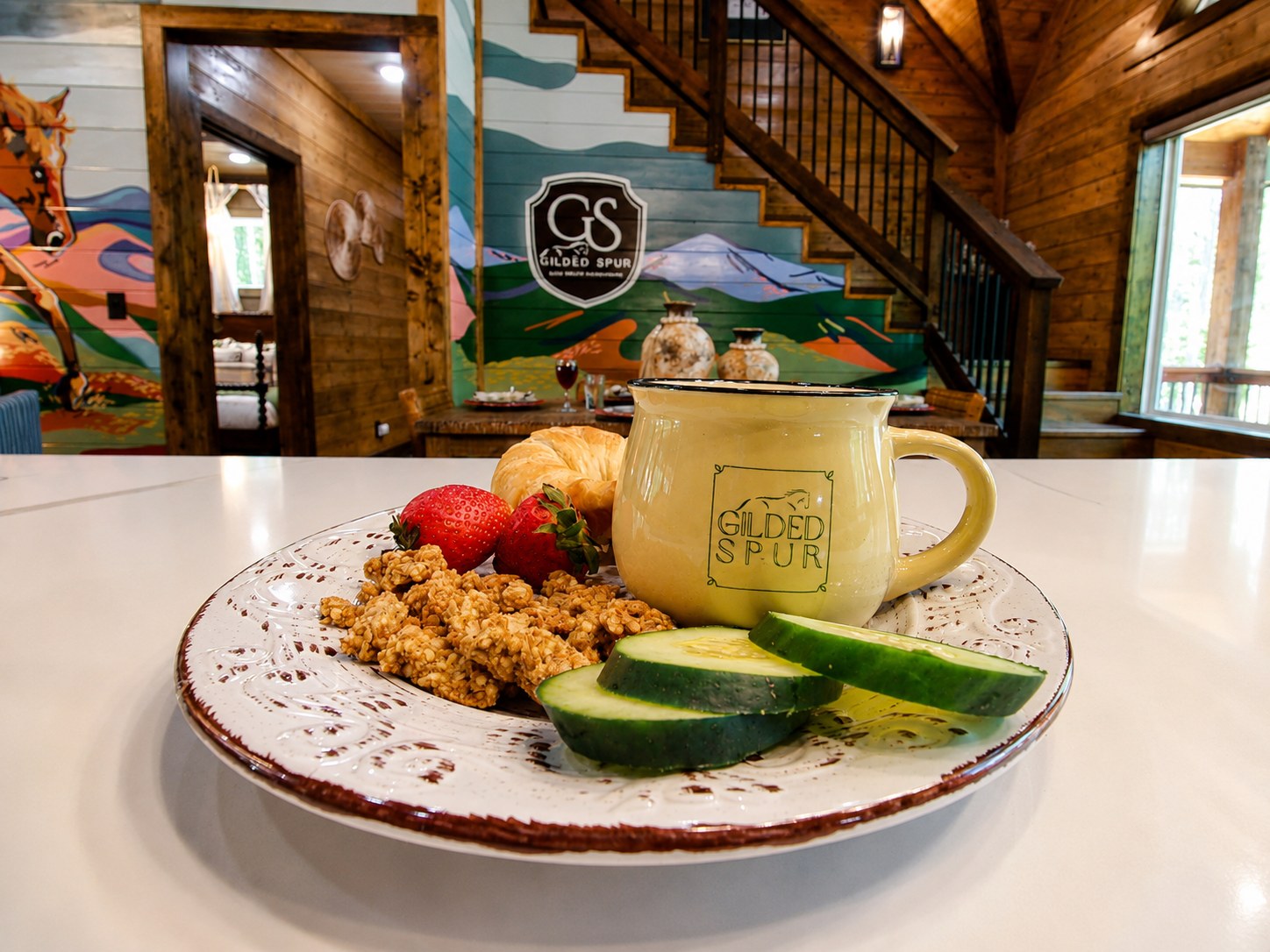 Gilded Spur branded ceramic plate and yellow mug at breakfast with granola, croissant, and fruit — GS crest signage on the mural wall behind, Hochatown Oklahoma equestrian-luxury cabin