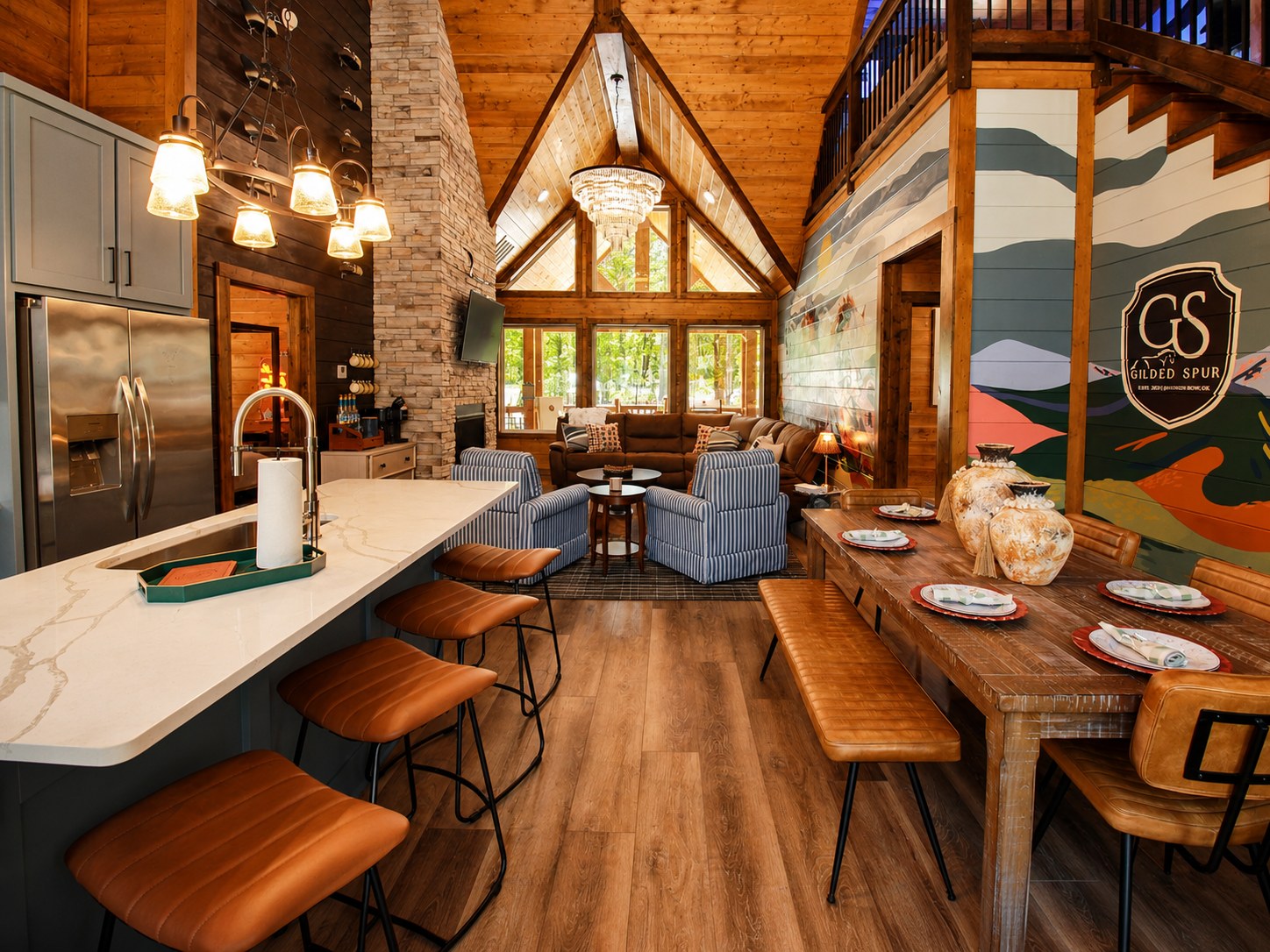 Gilded Spur cabin great room after Yellowwood Atelier design, Hochatown OK equestrian-luxury vacation rental: marble island, leather counter stools, brass chandelier, vintage duck decoys on stone chimney column, GS crest signage, horse mural visible