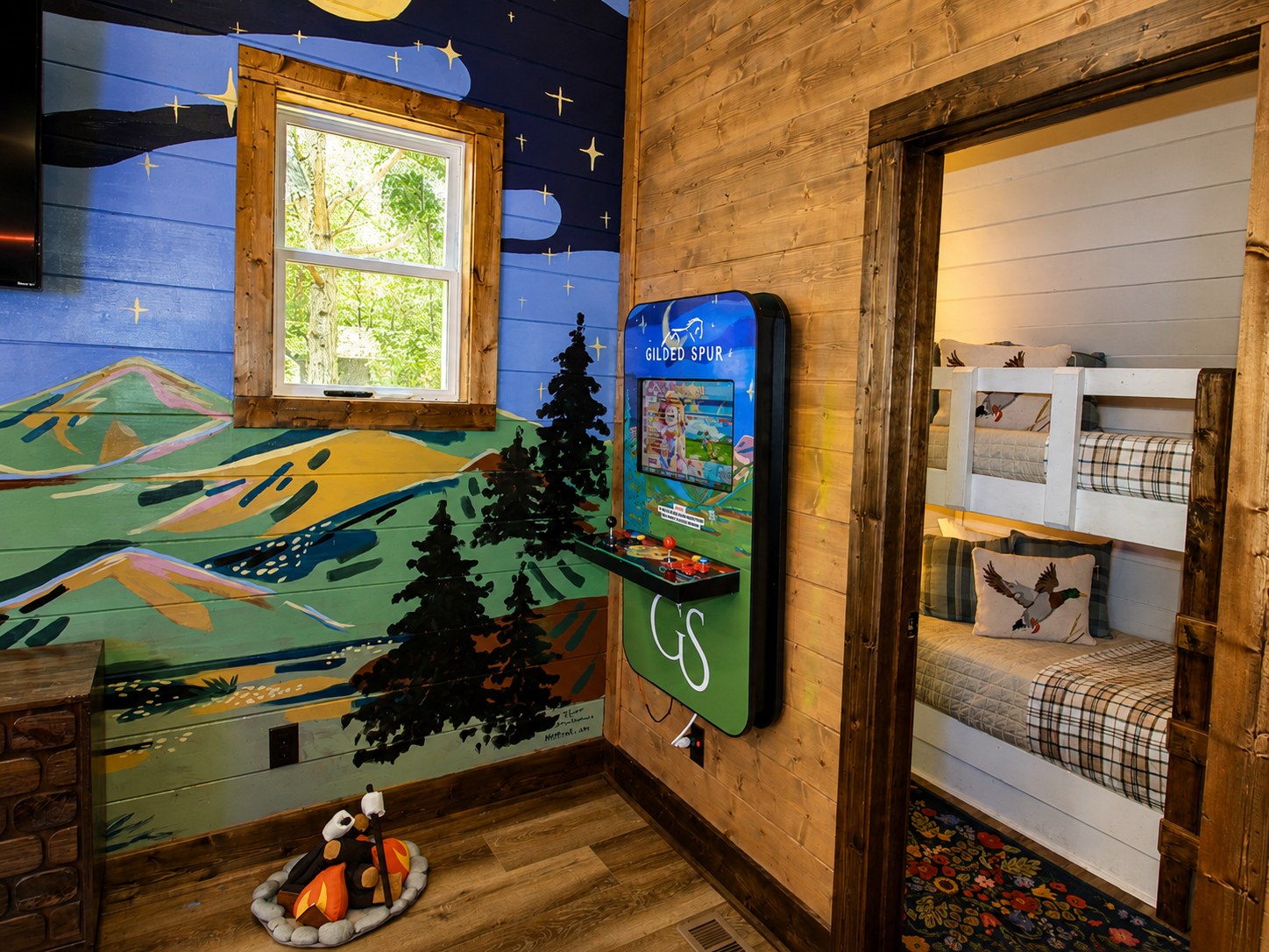 Gilded Spur upstairs loft game zone after Yellowwood Atelier design, Hochatown OK luxury vacation rental: full-size arcade, hand-painted Oklahoma landscape slant-wall mural, custom bunks, cowhide rug