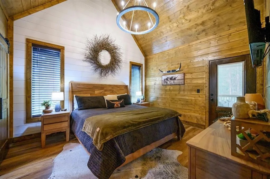 Gilded Spur primary bedroom before Yellowwood Atelier design, Broken Bow OK cabin: cathedral wood walls, sunburst chandelier, basic gray quilted bedding, barn door to bath
