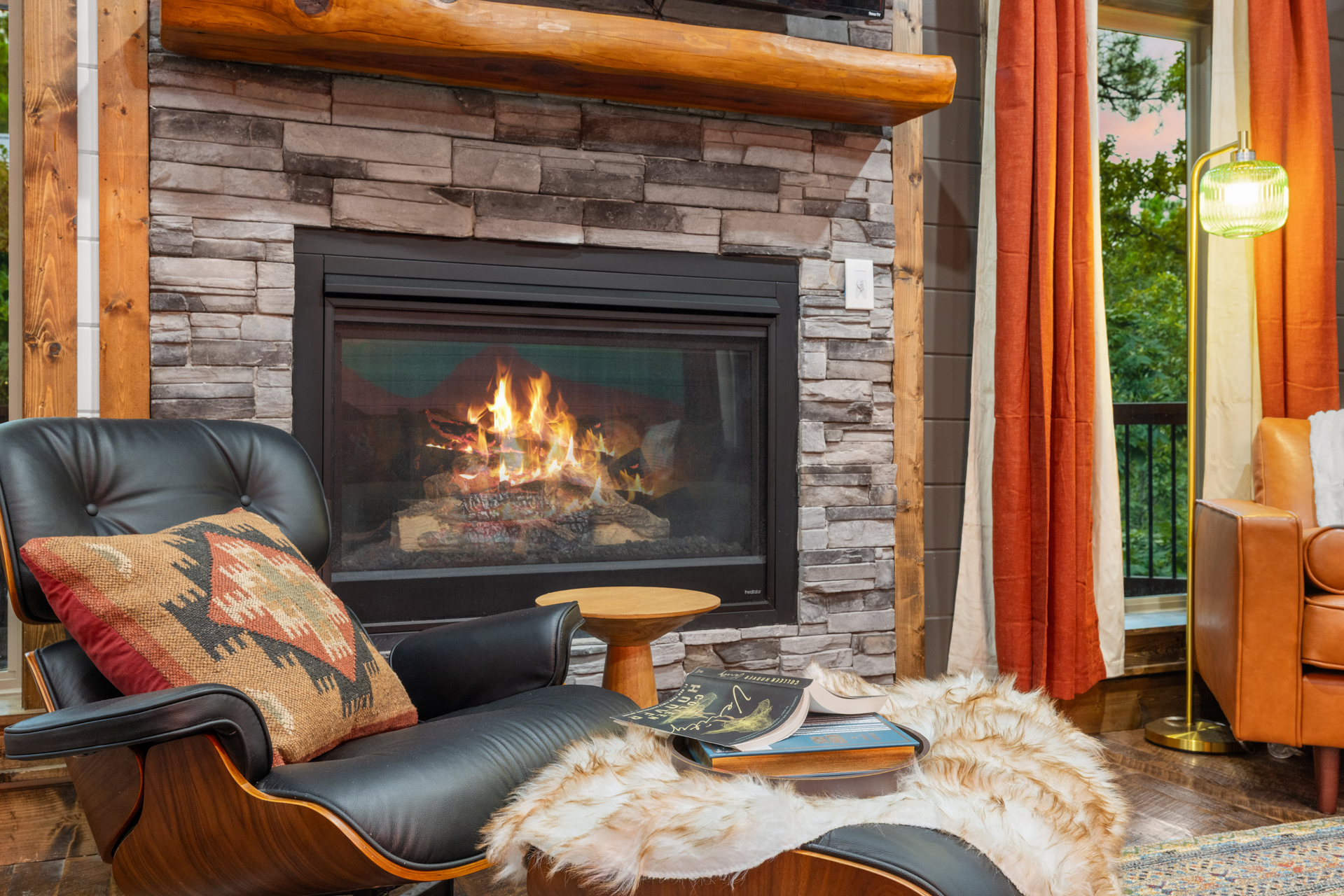 Eames lounge chair detail with red Aztec pillow, fur throw, books, brass lamp, beside the stone fireplace