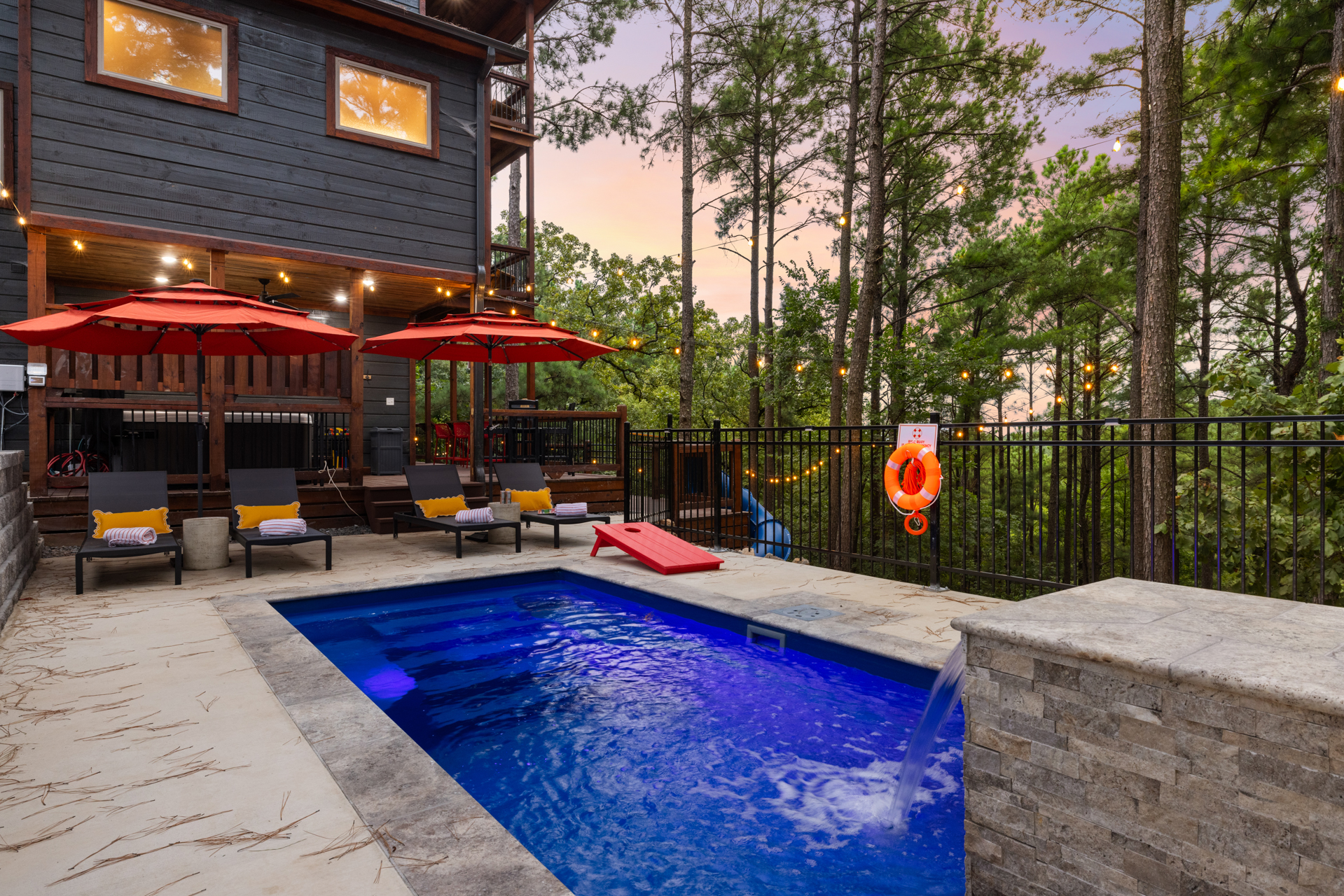 The Lazy Lariat pool deck at sunset with red umbrellas, cabin lit up, string lights, Broken Bow Oklahoma luxury cabin and pool short-term rental