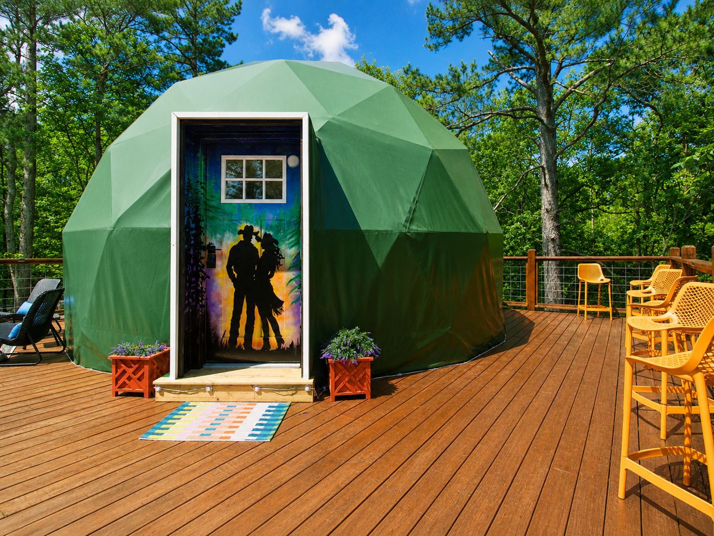 The Space Cowboy geodesic stargazing dome with a hand-painted cowboy-couple silhouette on the door, deck seating and forest beyond, Broken Bow Oklahoma glamping short-term rental