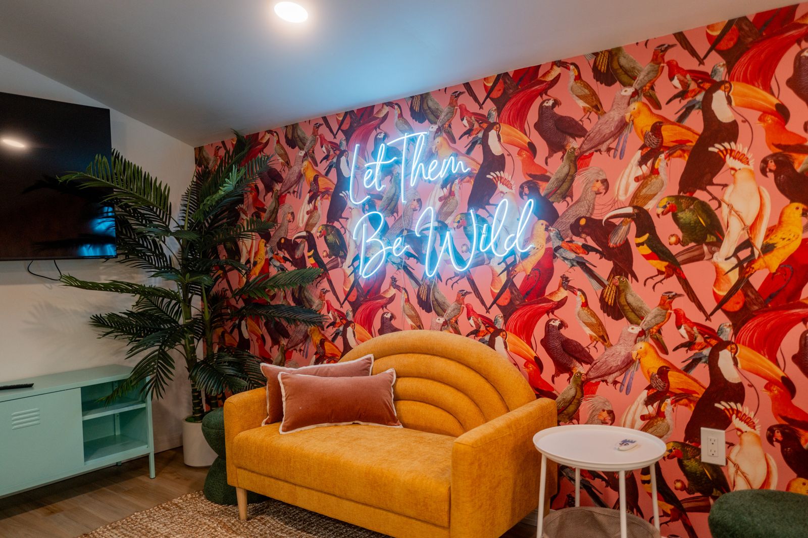 &lsquo;Let Them Be Wild&rsquo; neon over a mustard sofa and tropical-bird wallpaper at The Wild Pelican in Bradenton, Florida